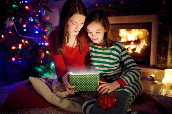 Szczęśliwa młoda matka i jej młoda córka otwierają magiczny prezent świąteczny przy kominku w przytulnym ciemnym salonie w Wigilię. — Zdjęcie stockowe