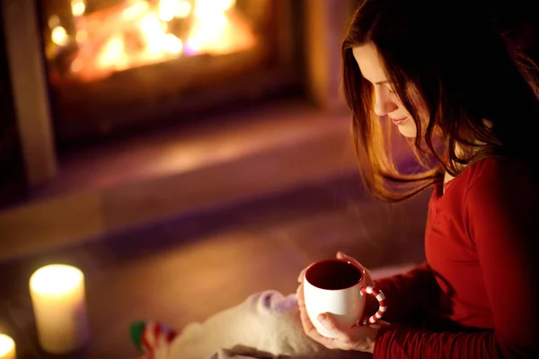 Happy young woman having a cup of hot chocolate by a fireplace in a cozy dark living room on Christmas eve. Celebrating Xmas at home. — Stock Photo, Image