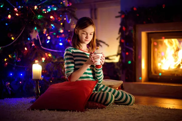Szczęśliwa młoda dziewczyna o filiżankę gorącej czekolady przy kominku w przytulnym ciemnym salonie w Wigilię. Świętowanie Bożego Narodzenia w domu. — Zdjęcie stockowe