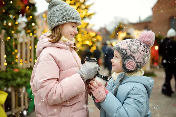 ラトビアのリガで伝統的なクリスマスフェアでホットチョコレートを飲む2人の愛らしい姉妹。クリスマスマーケットでお菓子やキャンディー、ジンジャーブレッドを楽しむ子供たち. — ストック写真