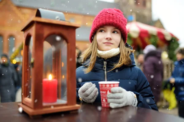 Roztomilá mladá dívka pije horkou čokoládu na tradičním vánočním veletrhu v Rize v Lotyšsku. Dítě se těší sladkosti, bonbóny a perník na vánočním trhu. — Stock fotografie