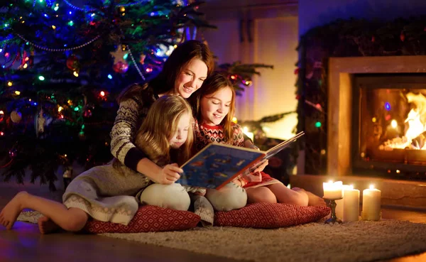 Feliz madre joven y sus hijas leyendo un libro de cuentos juntos junto a una chimenea en una acogedora sala de estar oscura en la víspera de Navidad. Celebrando la Navidad en casa . — Foto de Stock