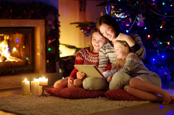 Mother and her two cute young daughters using a tablet pc at home by a fireplace in warm and cozy living room on Christmas eve. — Stock Photo, Image