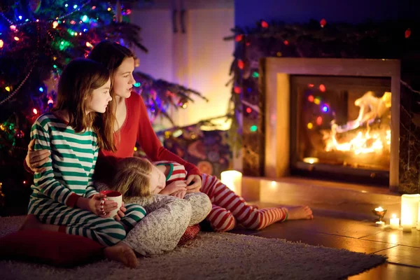 Happy young mother and her daughters having a good time sitting together by a fireplace in a cozy dark living room on Christmas eve. Celebrating Xmas at home.