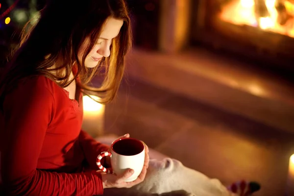 Vrolijke jonge vrouw met een kopje warme chocolademelk bij een open haard in een gezellige donkere woonkamer op kerstavond. Thuis Kerstmis vieren. — Stockfoto