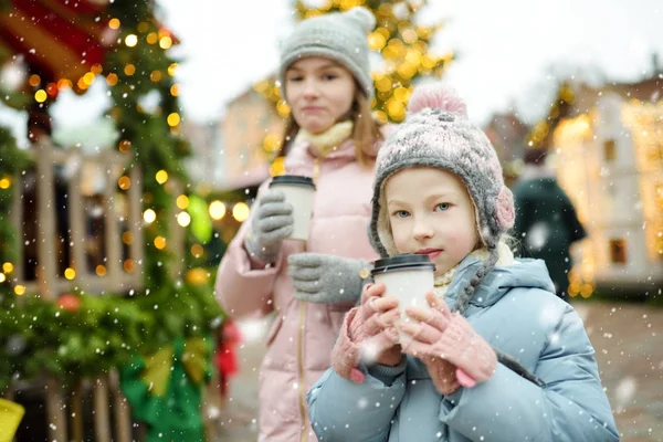 ラトビアのリガで伝統的なクリスマスフェアでホットチョコレートを飲む2人の愛らしい姉妹 クリスマスマーケットでお菓子やキャンディー ジンジャーブレッドを楽しむ子供たち 家族や子供と冬の時間 — ストック写真