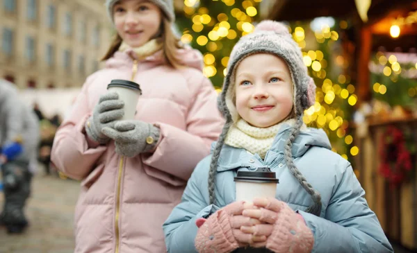 ラトビアのリガで伝統的なクリスマスフェアでホットチョコレートを飲む2人の愛らしい姉妹 クリスマスマーケットでお菓子やキャンディー ジンジャーブレッドを楽しむ子供たち 家族や子供と冬の時間 — ストック写真