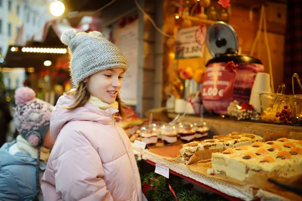 ラトビアのリガにある伝統的なクリスマスマーケットでお菓子を選ぶかわいい女の子 クリスマスにキャンディーとクッキーを買う子供 寒い冬の日の幸せな家族の時間 — ストック写真