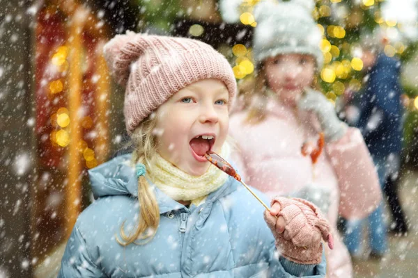 Twee schattige zusjes met hanenvormige lolly 's op de traditionele kerstmarkt in Riga, Letland. Kinderen die genieten van snoep, snoep en peperkoek op de kerstmarkt. — Stockfoto