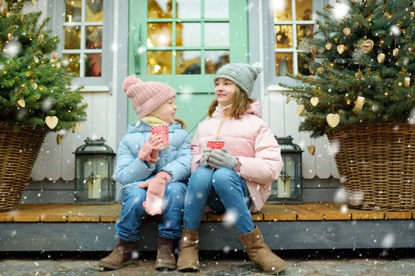 ラトビアのリガで伝統的なクリスマスフェアでホットチョコレートを飲む2人の愛らしい姉妹 クリスマスマーケットでお菓子やキャンディー ジンジャーブレッドを楽しむ子供たち 家族や子供と冬の時間 — ストック写真