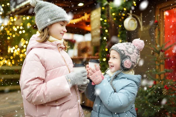 ラトビアのリガで伝統的なクリスマスフェアでホットチョコレートを飲む2人の愛らしい姉妹。クリスマスマーケットでお菓子やキャンディー、ジンジャーブレッドを楽しむ子供たち. — ストック写真