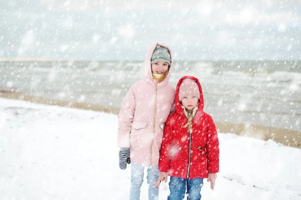 Δύο χαριτωμένες μικρές αδελφές διασκεδάζουν μαζί στη χειμερινή παραλία την κρύα χειμωνιάτικη μέρα. Παιδιά που παίζουν δίπλα στον ωκεανό. — Φωτογραφία Αρχείου