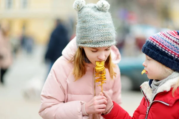 Cute little girls eating fried potato on a stick on cold winter day outdoors. — Stock Photo, Image