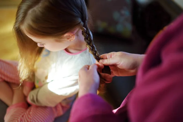 Mor flätar sitt lilla dotters hår på morgonen. Söt liten flicka gör sig redo för skolan. — Stockfoto