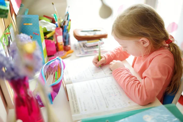 Smart liten skolflicka som gör sina läxor vid bordet hemma. Barn lär sig att skriva och läsa. — Stockfoto