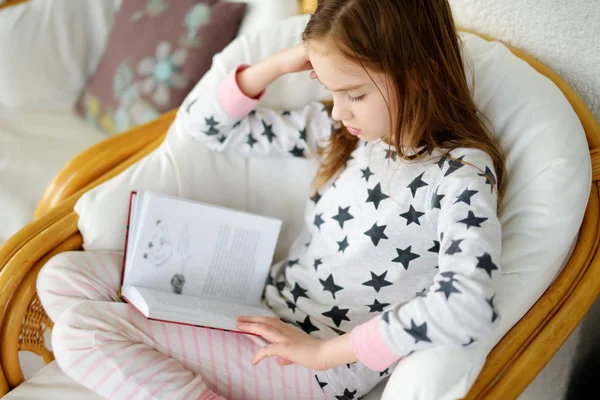 Menina adorável lendo um livro na sala de estar branca no belo dia de verão. Estudante inteligente fazendo seu dever de casa em casa depois da escola . — Fotografia de Stock