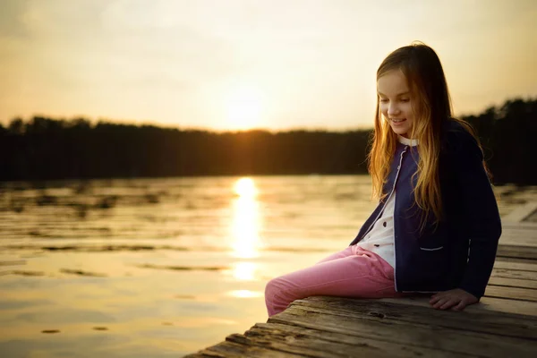 Linda chica sentada en una plataforma de madera junto al río o lago sumergiendo sus pies en el agua en el cálido día de verano. Actividades familiares en verano . — Foto de Stock