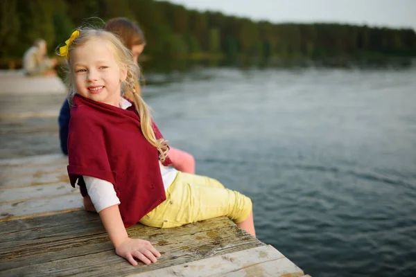 Due ragazze carine sedute su una piattaforma di legno vicino al fiume o al lago immergendo i piedi in acqua nelle calde giornate estive. Attività per famiglie in estate . — Foto Stock