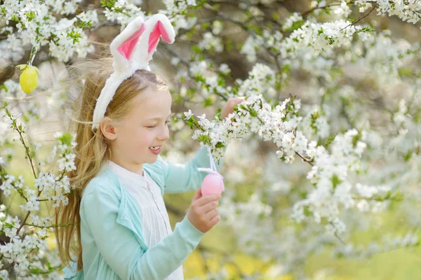 美しい春の日に桜の庭にウサギの耳を身に着けている愛らしい少女 桜の枝にイースターエッグをぶら下げている子供 屋外でイースターを祝う子供 — ストック写真