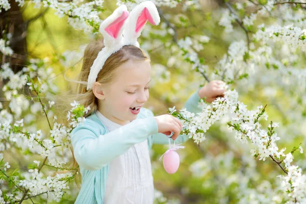 美しい春の日に桜の庭にウサギの耳を身に着けている愛らしい少女 桜の枝にイースターエッグをぶら下げている子供 屋外でイースターを祝う子供 — ストック写真