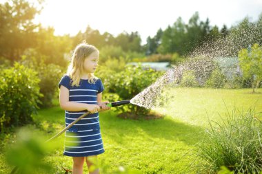 Cute young girl watering flower beds in the garden at summer day. Child using garden hose on sunny day. Kid helping with everyday chores. Mommys little helper. clipart
