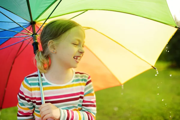 夏の雨の日にカラフルな虹の傘を持ってかわいい女の子 屋外で暖かい雨の下を歩く子供 子供のための屋外夏の活動 — ストック写真