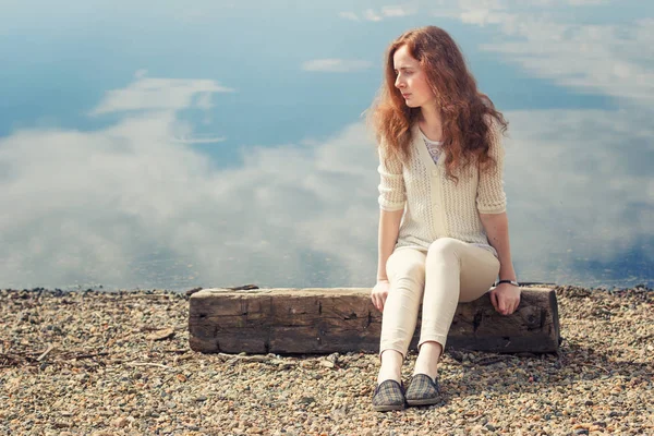 The red-haired young woman sits alone on the shore on a summer day. — Stock Photo, Image