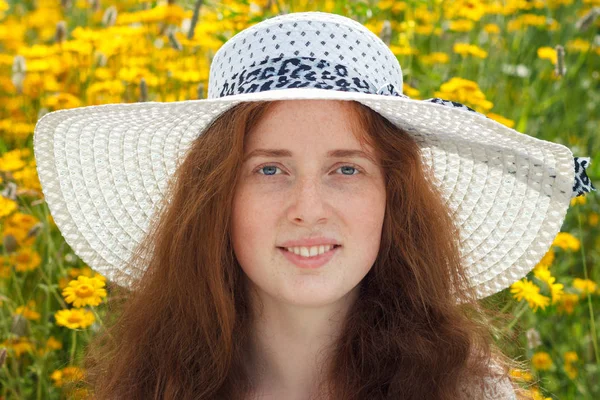 Portret van roodharige jonge vrouw in witte hoed op een achtergrond van zomerbloemen. — Stockfoto