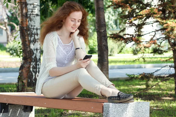 Jovem feliz com telefone celular sentado em um banco em um parque da cidade . — Fotografia de Stock