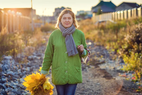 秋の葉の花束を持つ幸せな女性が秋の日に村の通りを歩いています — ストック写真