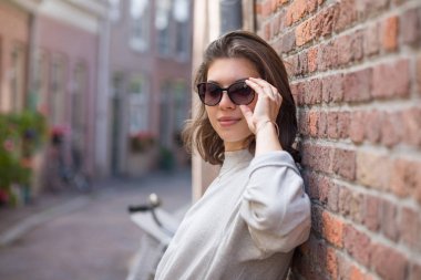 Güneş gözlüklü mutlu bir genç kadın, yaz mevsiminde şehir sokağında duvara yaslanmış duruyor. Modern sokak modası..