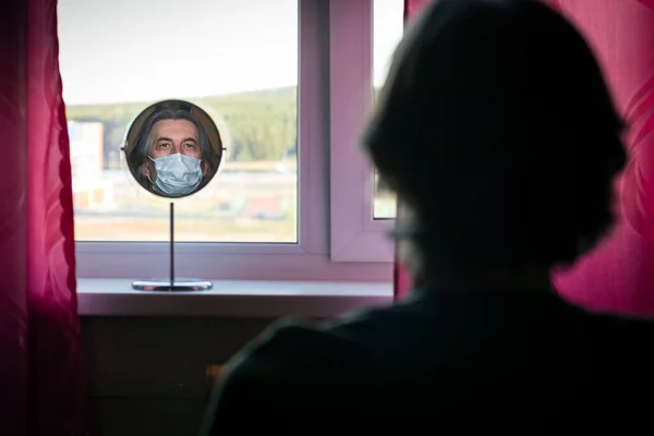 Reflection of a portrait of a man in a medical mask in a round mirror standing on the windowsill of the apartment. The concept of isolation of people during coronaviruses infection.