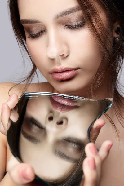 Female with mirror shard in hand posing on gray background.