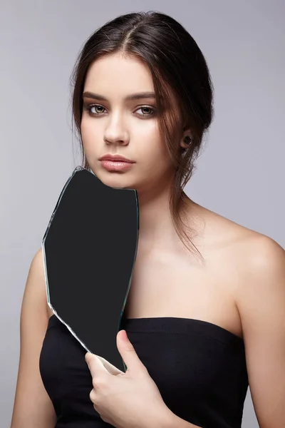 Female with mirror shard in hand posing on gray background.