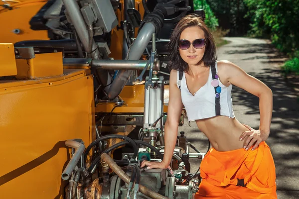 Sexig kvinna arbetare i overaller står nära traktorn. — Stockfoto
