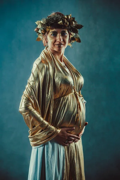 Schwangere in goldener Toga und Kranz posiert wie eine griechische Fruchtbarkeitsgöttin — Stockfoto