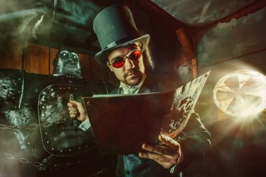 Genç sakallı adam silindir şapka büyük robot yakın dururken el ile kitap okuyor.