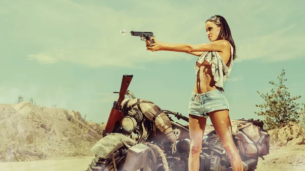 Ποδηλάτης σέξι γυναίκα με το όπλο κοντά μηχανάκι στο έρημο φόντο. — Φωτογραφία Αρχείου