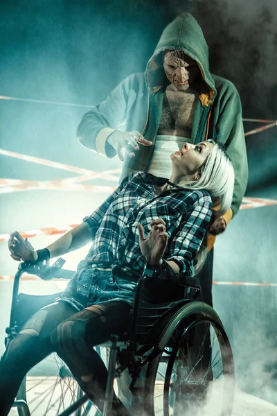 Maniaco e la sua vittima legata sulla sedia a rotelle sullo sfondo scuro . — Foto Stock