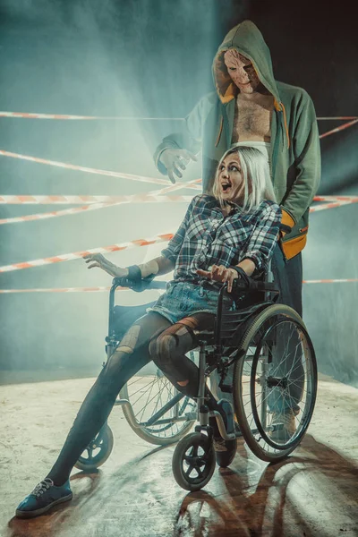 Maniac y su víctima atada en la silla de ruedas en el fondo oscuro . — Foto de Stock