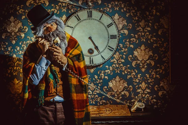 Uomo anziano caucasico in piedi vicino orologio da parete nella stanza buia e parlando al telefono . — Foto Stock
