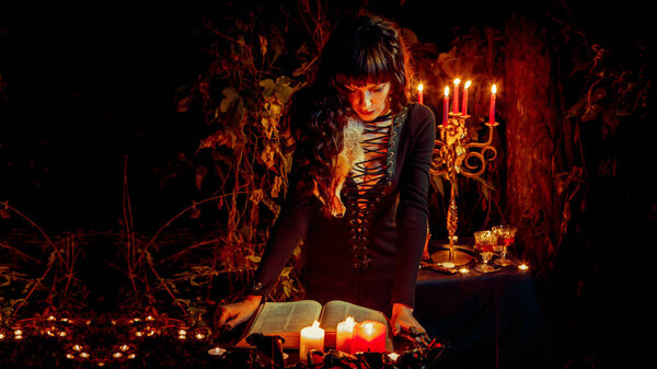 Beautiful witch is reading the vintage book of magic on the forest background.