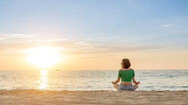 Beyaz kadın deniz kıyısında yoga yapıyor. Arkaplanda gün batımı.