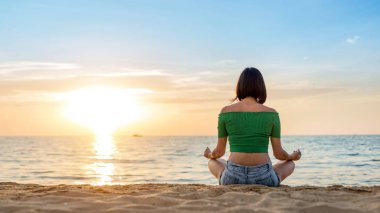 Beyaz kadın deniz kıyısında yoga yapıyor. Arkaplanda gün batımı.