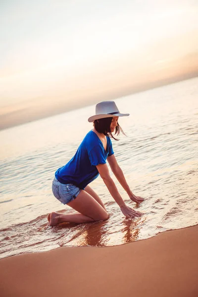 Mujer joven en sombrero está tocando el agua en el fondo de la puesta del sol . — Foto de Stock