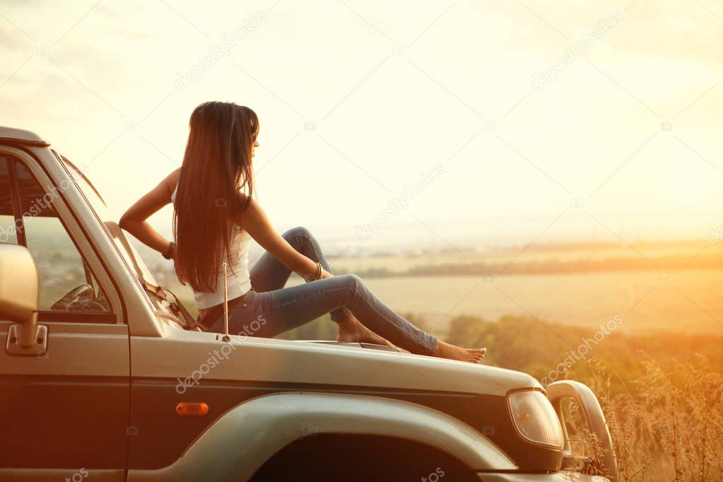 Attractive yong woman is sitting on the car's hood and looking at sunset. Rural evening background.