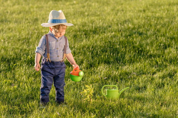Portrait of toddler child outdoors. Rural scene with one year old baby boy wearing straw hat using watering can