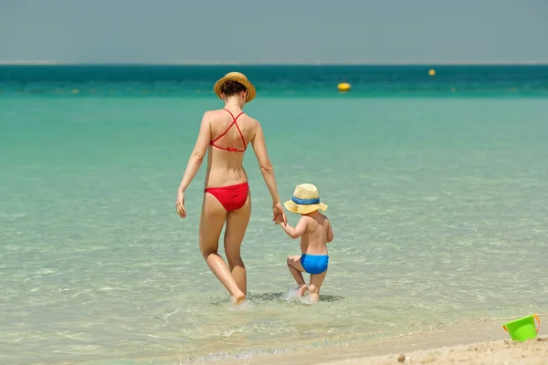 きれいなブルーの水とビーチで帽子で母親と一緒に歩いているかわいい幼児少年 — ストック写真