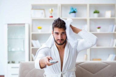 Bornozlu genç adam evde kanepede televizyon izliyor.