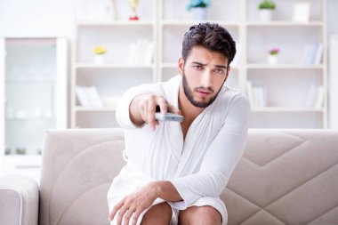 Bornozlu genç adam evde kanepede televizyon izliyor.